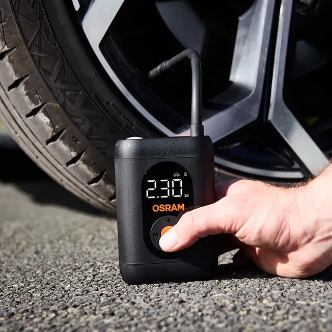 Hånd trykker på tænd/sluk-knap på Osram TYREinflate 1600 ved bildæk, display viser 230 kPa