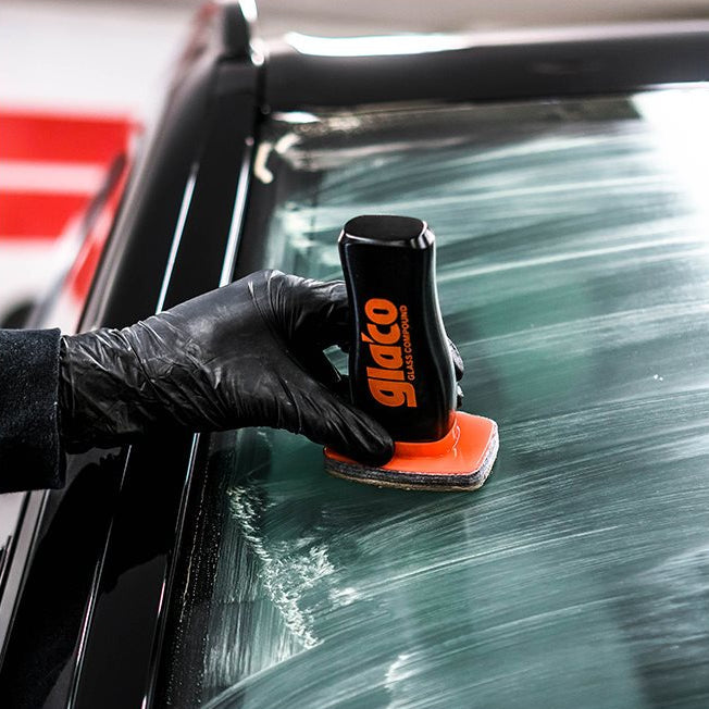 Een hand met een zwarte handschoen brengt Glaco Glass Compound aan op een autoruit.